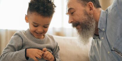 A young child, with curly dark hair, is seated on the couch next to his grandfather, who has a long beard and is wearing a gray sweater. His grandfather watches over him as he plays with his toy cars.