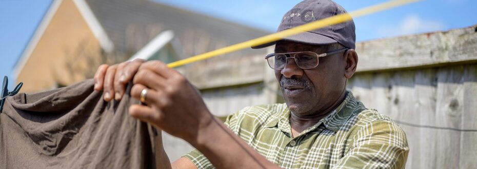 An elderly black man with glasses, a dark blue cap and wearing a green and white checkered shirt, pins his clothing to a washing line to dry out in the sun.