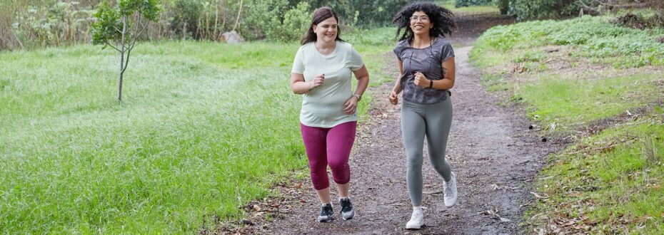 Two women running together on a dirt path in a park, one white middle-aged woman is wearing a white t-shirt and pink leggings, while the other young black woman is wearing a grey t-shirt and grey leggings.