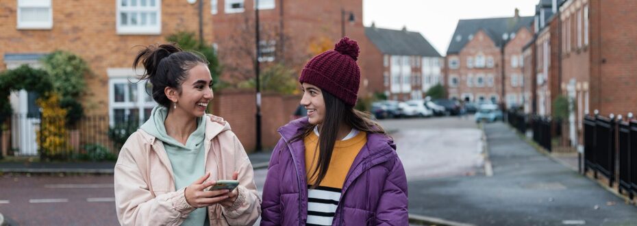 Two smiling sisters walking and chatting together on a residential street, in a city neighbourhood. One of the sisters is wearing a mint green hoodie and holding a phone, while the other has on a purple puffy coat, yellow top, blue jeans and a maroon knit hat with a pom-pom on top, laughing with eachother.