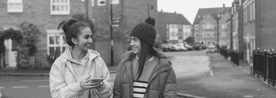 Two smiling sisters walking and chatting together on a residential street, in a city neighbourhood. One of the sisters is wearing a mint green hoodie and holding a phone, while the other has on a purple puffy coat, yellow top, blue jeans and a maroon knit hat with a pom-pom on top, laughing with eachother.