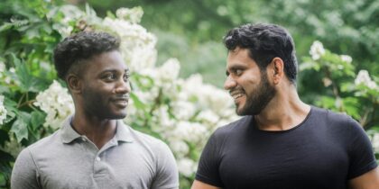 Two men standing outdoors on a walk. One Black man is wearing a grey shirt, and the other South Asian man is wearing a black shirt. They are both smiling at eachother and looking lovingly.