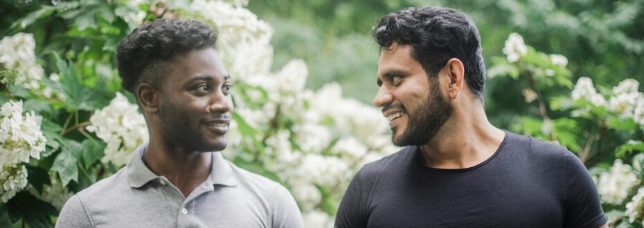 Two men standing outdoors on a walk. One Black man is wearing a grey shirt, and the other South Asian man is wearing a black shirt. They are both smiling at eachother and looking lovingly.
