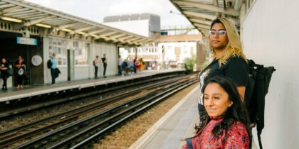 Two women are stood standing waiting for a train at the platform. One South Asian woman with dark brown and blonde hair is wearing glasses, a black shirt and a bag pack, while the other South Asian woman with dark brown hair is wearing a bright red, printed dress.