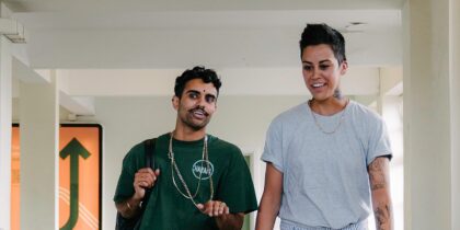 A young South Asian man wearing a green t-shirt and a chain necklace is having a conversation with a young South Asian woman with short dark hair and tattoos on her arm and neck wearing a grey t-shirt and a gold necklace. The pair appear to be laughing while walking down a hallway.