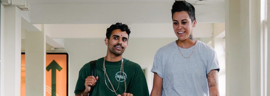 A young South Asian man wearing a green t-shirt and a chain necklace is having a conversation with a young South Asian woman with short dark hair and tattoos on her arm and neck wearing a grey t-shirt and a gold necklace. The pair appear to be laughing while walking down a hallway.
