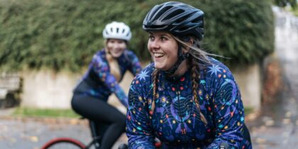 Two smiling women wearing bicycle helmets and dressed in colorful patterned outfits. They're both outdoors, perhaps on a cycling trail or path surrounded by greenery. The woman in the foreground is wearing a vibrant blue jacket with stars and floral patterns, and with two long plaited braids. She is seated on a bicycle, looking directly at the camera with a joyful expression.