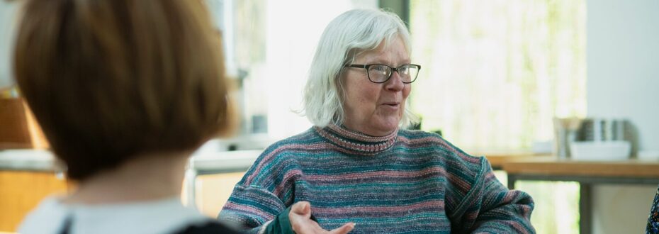 An elderly white woman with grey hair and glasses engaged in conversation, holding a mug. She is wearing a colorful striped sweater and gesturing with her hand while speaking. She is talking to a group of friends.