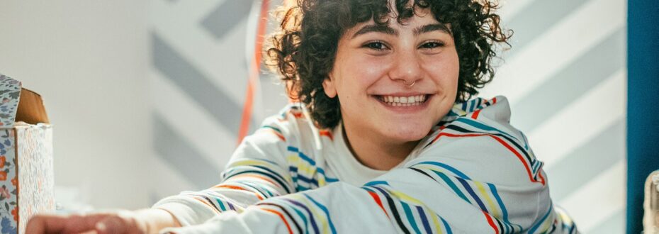 A young person with curly brown hair, wearing a colourful striped shirt, is smiling directly to the camera.