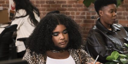 Two colleagues of varying genders are sat at a desk working. One colleague has a curly dark, and is wearing a leopard print jacket over a white top and a chunky chain necklace. The colleague next to them is wearing a dark brown leather jacket. They both have a pensive expression.