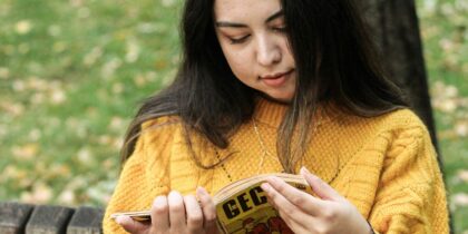 A young white woman with long brunette hair, wearing a yellow sweater is sitting outdoors in a park on a bench, reading a book.