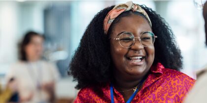 A Black woman with curly hair wearing round glasses, a colorful patterned headband, and a bright red and pink leopard print top is smiling while in conversation with work colleagues.