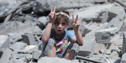 A young girl sits among rubble and debris, in Gaza. She has curly brown hair with a brown and grey stripped hairband, and wears a colourful floral patterned outfit, likely pyjamas. She's looking directly to camera with holding up a peace sign.