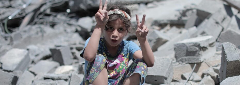 A young girl sits among rubble and debris, in Gaza. She has curly brown hair with a brown and grey stripped hairband, and wears a colourful floral patterned outfit, likely pyjamas. She's looking directly to camera with holding up a peace sign.