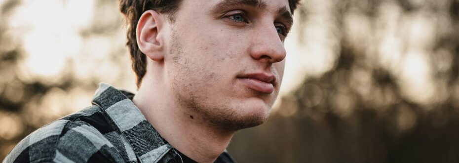 A close-up profile view of a young white man with short dark brown hair, sitting outdoors by some trees. He has blue eyes and is wearing a black and grey plaid flannel shirt.