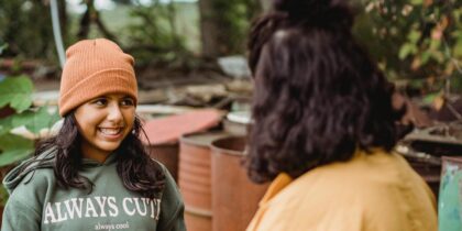 A young Asian girl is smiling at her Mother, she has long brown hair and is wearing an orange beanie and green hoodie reading 'Always cute'. They appear to be in a garden together. Her Mother has short brown hair and a mustard shirt.