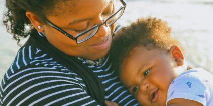 Close up of a Black mother with glasses and an afro holding a smiling baby. The Mother is wearing a black and white striped shirt and is looking down at the baby, who is wearing a white onesie. The pair are outdoors, by a beach.