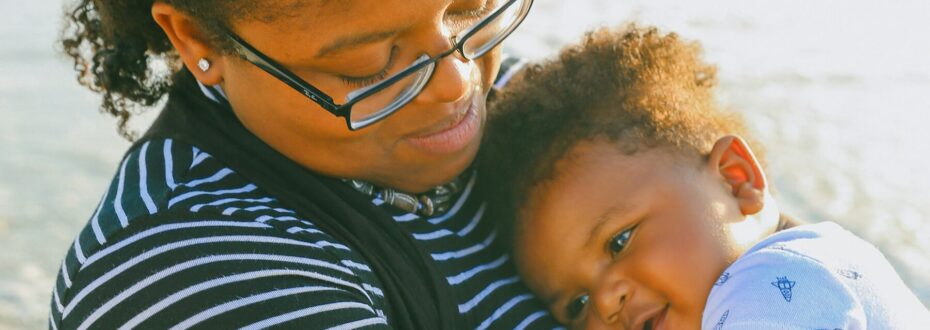 Close up of a Black mother with glasses and an afro holding a smiling baby. The Mother is wearing a black and white striped shirt and is looking down at the baby, who is wearing a white onesie. The pair are outdoors, by a beach.