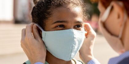 Young mixed race girl is looking at a parent who is helping them put their face mask on during Covid-19.