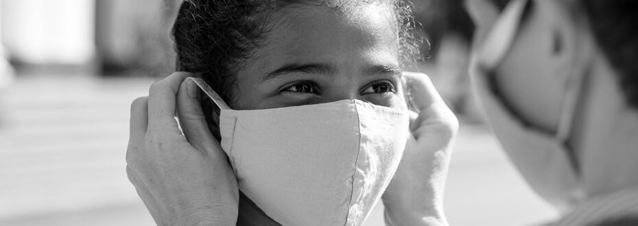 Young mixed race girl is looking at a parent who is helping them put their face mask on during Covid-19.