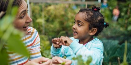 A mother and her daughter are gardening together in an allotment. The Mother is wearing a colourful striped shirt and is smiling at her daughter. The daughter is wearing a light blue top with her hair in three small buns and is smiling back at her Mother.