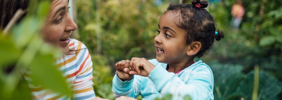 A mother and her daughter are gardening together in an allotment. The Mother is wearing a colourful striped shirt and is smiling at her daughter. The daughter is wearing a light blue top with her hair in three small buns and is smiling back at her Mother.