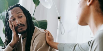 A Black man with dreadlocks is sat on a couch, confiding in a friend who is comforting him with her hand on his shoulder.