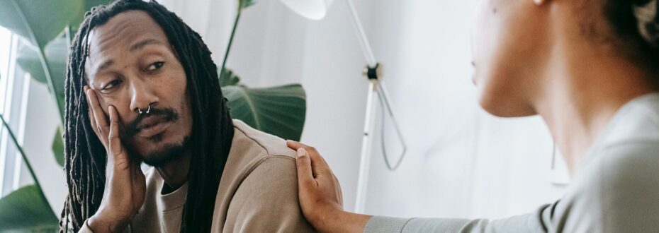 A Black man with dreadlocks is sat on a couch, confiding in a friend who is comforting him with her hand on his shoulder.