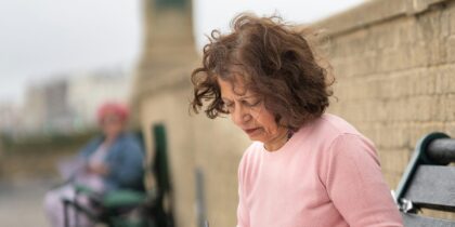 Older woman with brown hair wearing a pink top sits on bench outside, looking down.