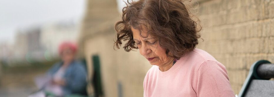 Older woman with brown hair wearing a pink top sits on bench outside, looking down.