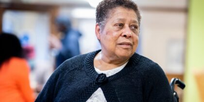 Older Black woman with short hair looks to one side. She is in a wheelchair and wearing a navy cardigan.