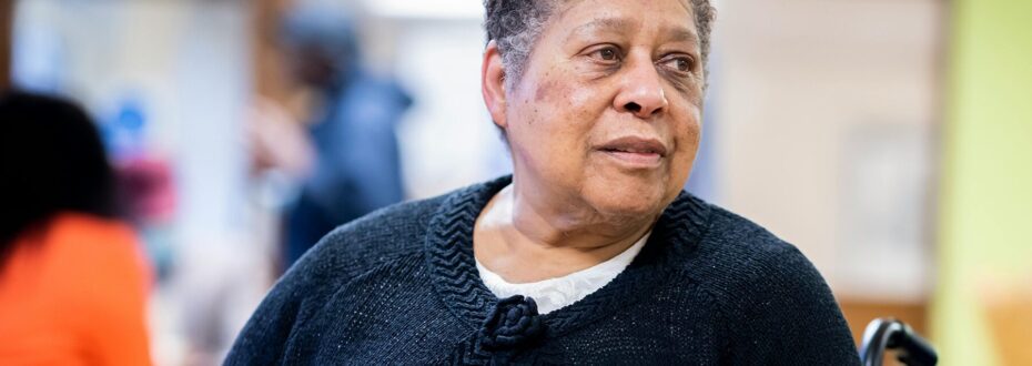 Older Black woman with short hair looks to one side. She is in a wheelchair and wearing a navy cardigan.