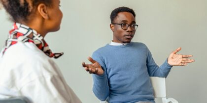 Young Black man wearing a sky-blue jumper in conversation with others, holding his hands out as he makes a point. Blurred in the foreground is a Black woman, wearing a white shirt, facing the man.