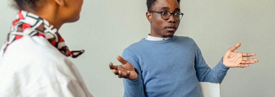 Young Black man wearing a sky-blue jumper in conversation with others, holding his hands out as he makes a point. Blurred in the foreground is a Black woman, wearing a white shirt, facing the man.