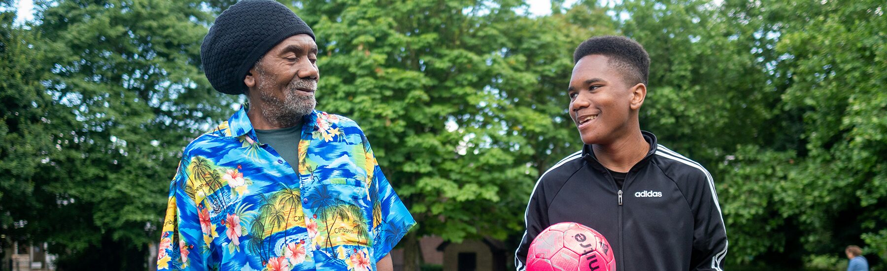 One older Black man and one teenage Black boy walking in a park together, one holds a red football