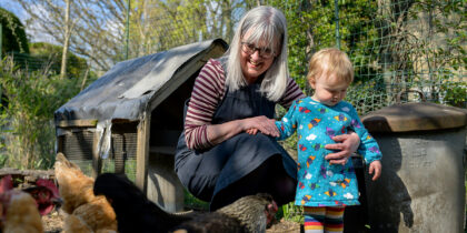 Older woman squats next to a toddler in a blue dress, feeding chickens