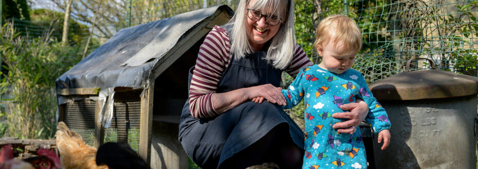 Older woman squats next to a toddler in a blue dress, feeding chickens