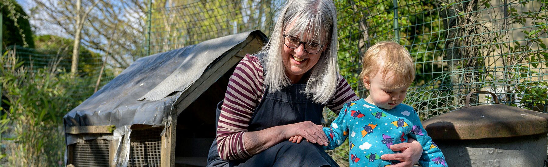 Older white woman with grey hair is squatting next to her toddler granddaughter in a garden