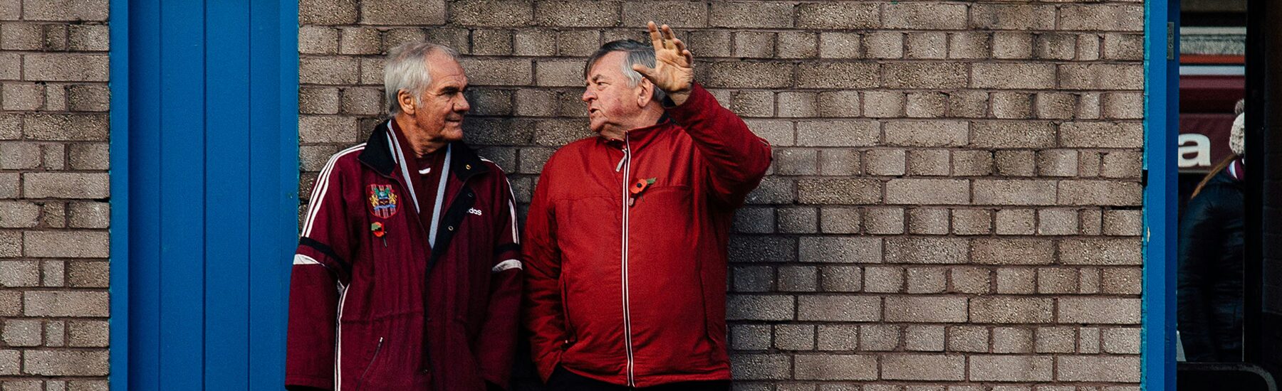 Two older white men stand outside against a brick wall, talking to each other