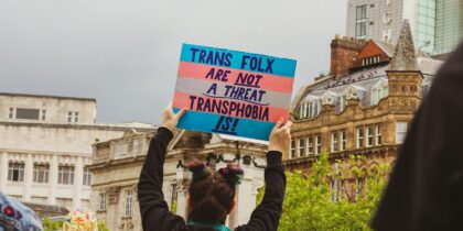 Person walking along in a crowd holds banner which reads: Trans folx are not a threat - transphobia is!